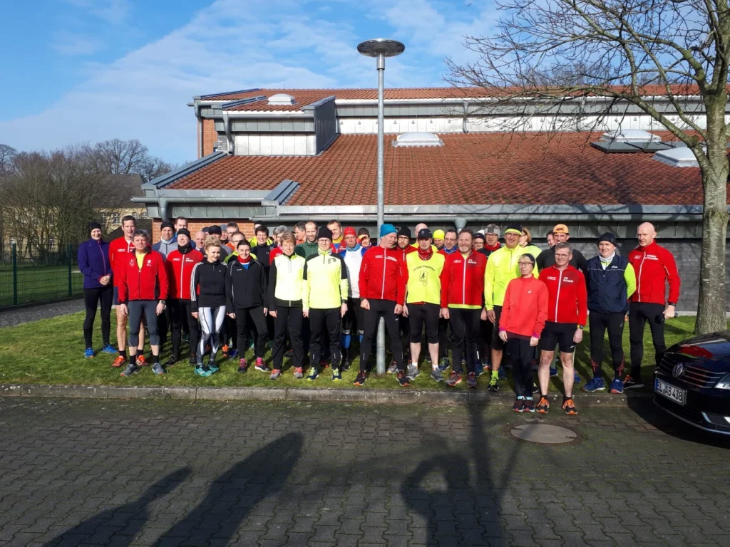 Gruppe von Läufern in bunter Sportkleidung posiert vor einem Gebäude bei sonnigem Wetter.