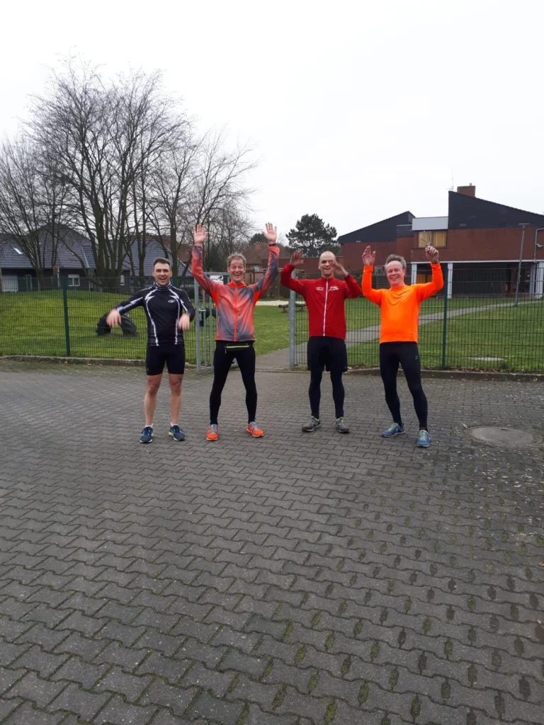 Vier Personen in Sportkleidung machen draußen Sprungübungen auf einem gepflasterten Platz vor einem Zaun.