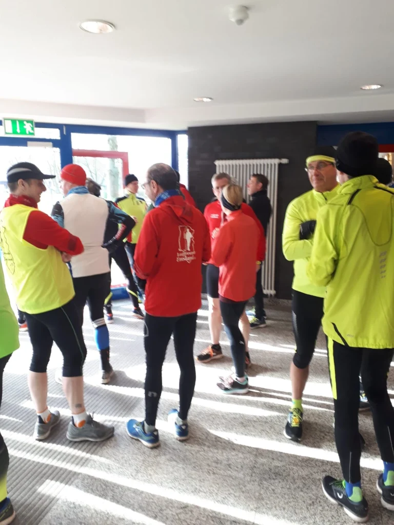 Gruppe von Läufern in Sportkleidung, die sich in einer Halle unterhalten. Einige tragen leuchtend gelbe und rote Jacken, während andere in schwarzer Sportkleidung gekleidet sind. Tageslicht fällt durch die Fenster und schafft eine freundliche Atmosphäre für das bevorstehende Lauf-Event.