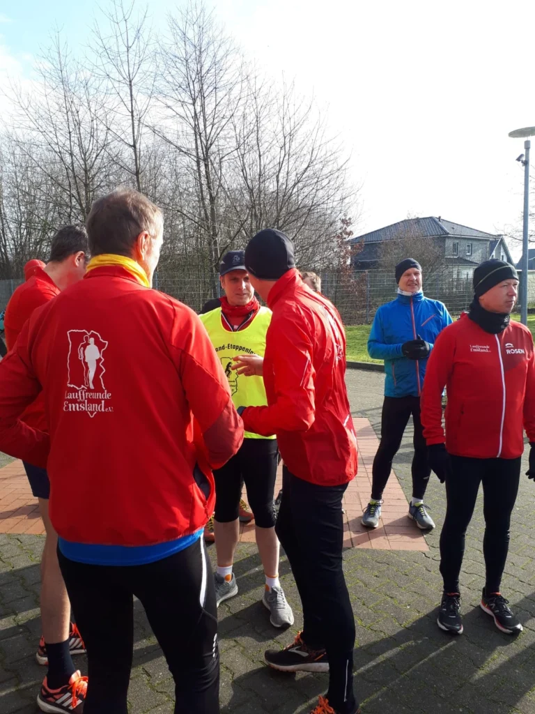 Gruppe von Joggern in roten Jacken bei sonnigem Wetter versammelt auf einem Platz mit Häusern und Bäumen im Hintergrund.