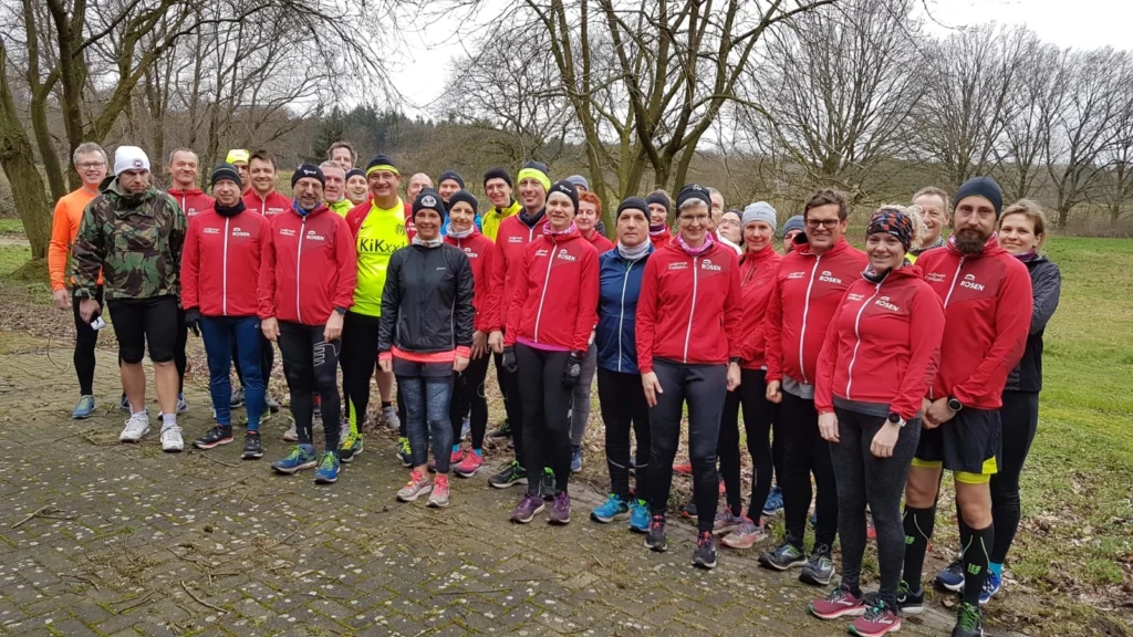 Gruppe von Läufern in roten Jacken posiert im Freien bei einem Parklauf; kaltes Wetter, kahlköpfige Bäume im Hintergrund.