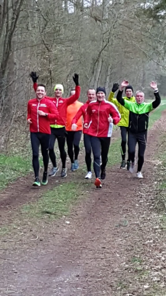 Gruppe von sieben Läufern in sportlicher Kleidung, die auf einem Waldweg joggen. Die Teilnehmer tragen rote, grüne und orange Laufjacken und winken fröhlich in die Kamera.