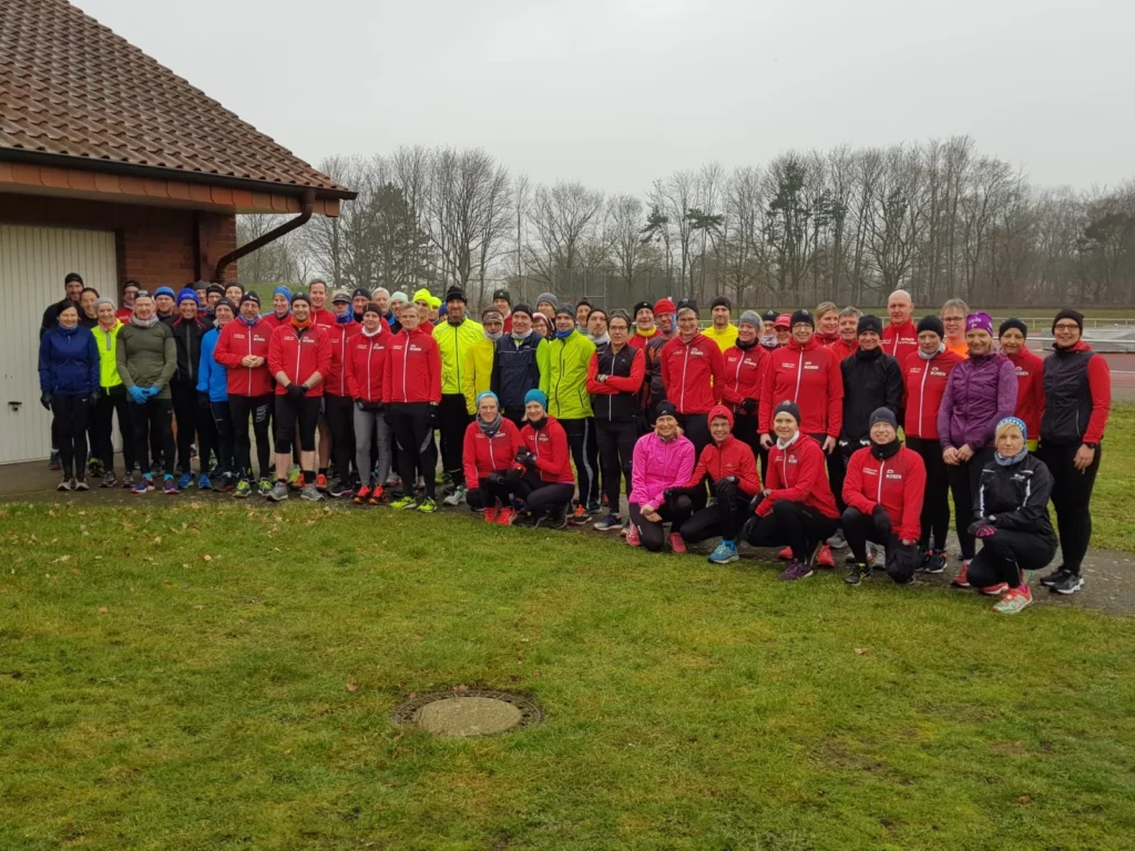 Gruppenfoto von Läufern in roten Jacken, die sich auf einem Sportgelände versammeln. Die Teilnehmer sind in verschiedenen Laufoutfits gekleidet und stehen vor einem Gebäude sowie einer Laufbahn, umgeben von Bäumen und Gras.