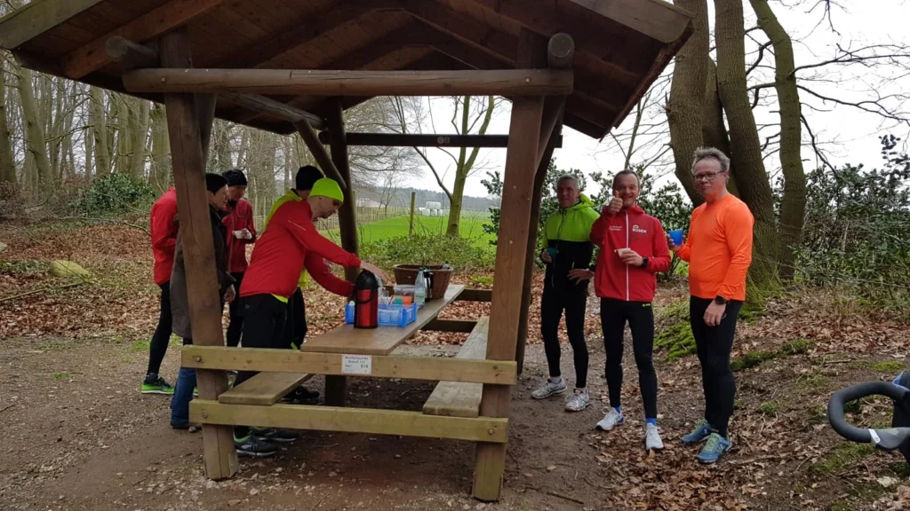 Läufergruppe beim Verpflegungstisch im Wald während einer Pause beim Training, mit Getränken und Snacks.
