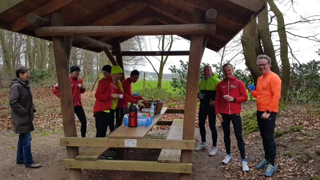 Läufer-Gruppe erholt sich im Wald unter einer Holzhütte mit Getränken auf einem Tisch.