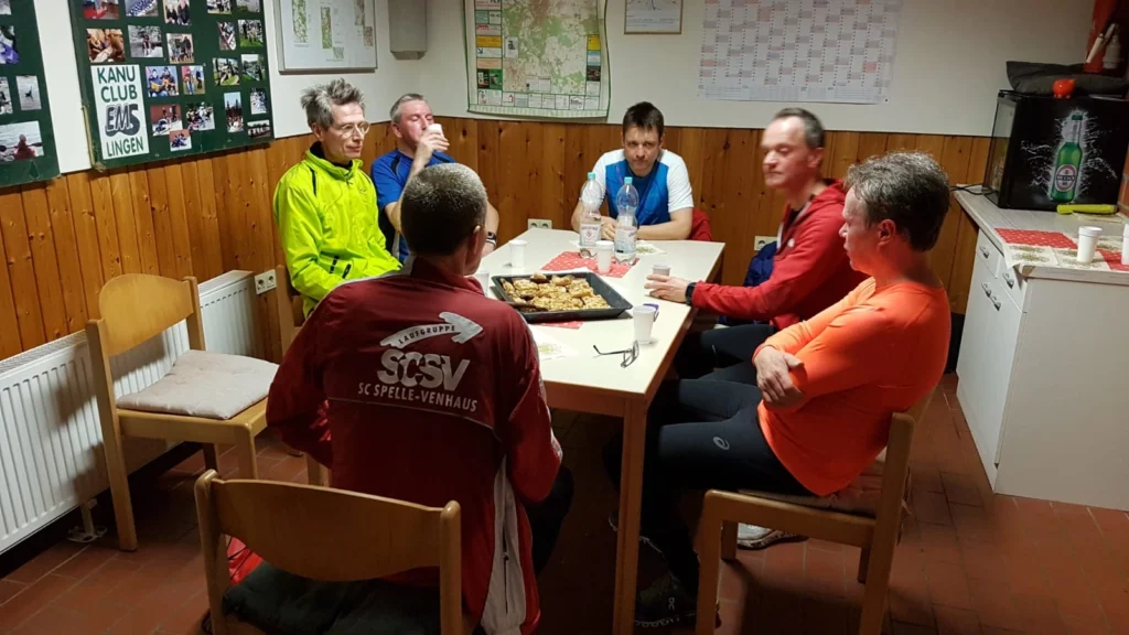 Gruppe von sechs Personen in einem Vereinsraum des Kanu Clubs EMS Lingen, sitzend am Tisch mit Snacks.