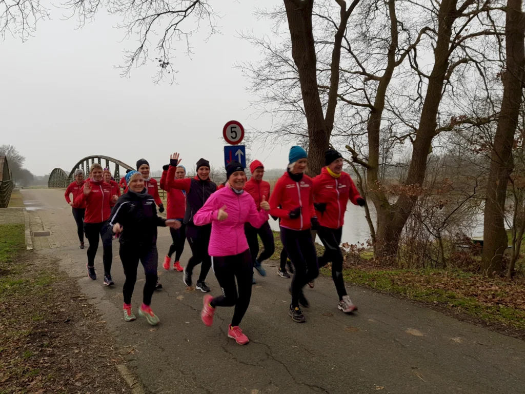 Gruppe von Läuferinnen in sportlicher Kleidung, die auf einem Weg entlang eines Flusses joggen. Sie tragen rote Jacken und lächeln, während sie an einem Brückenbogen vorbeilaufen. Der Himmel ist bewölkt, und die Umgebung ist von Bäumen gesäumt.