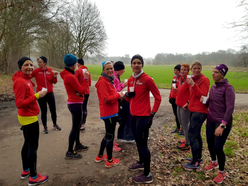 Läufergruppe in roten Jacken macht Pause auf Waldweg im Winter, grünes Feld im Hintergrund, relaxen beim Training.