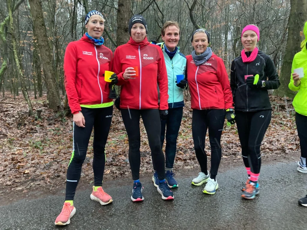 Gruppe von fünf Läuferinnen in sportlicher Kleidung, die in einem Wald stehen. Sie tragen rote Jacken und halten bunte Tassen. Der Boden ist nass und es sind vereinzelt Blätter zu sehen.