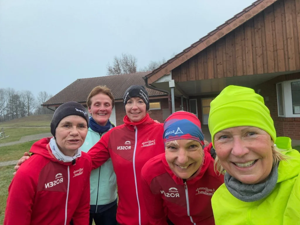Gruppe lächelnder Menschen in Sportkleidung vor einem Haus im Freien an einem bewölkten Tag.