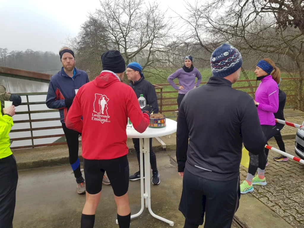 Gruppe von Läufern beim Austausch nach dem Training am Wasser, umgeben von Bäumen und einer Brücke. Einige Teilnehmer tragen Sportkleidung und Mützen, während sie Getränke und Snacks genießen.