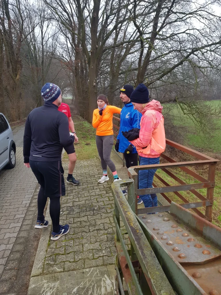 Gruppe von fünf Menschen im Freien, warm gekleidet, beim Gespräch nach dem Winterlauf im Park.