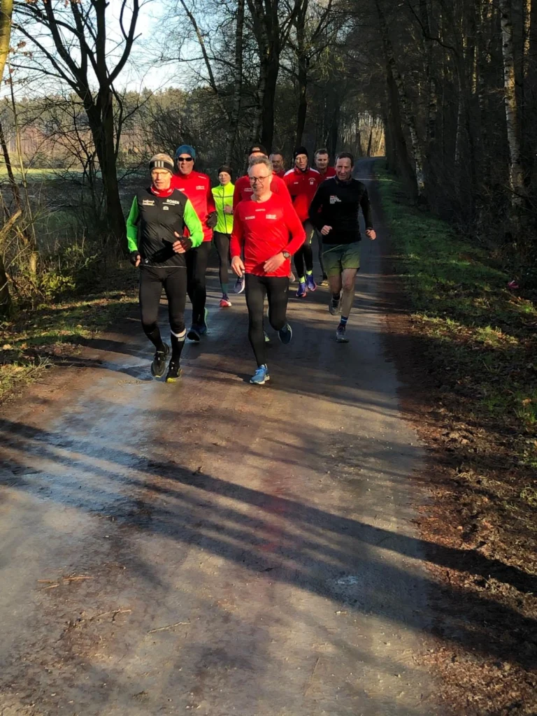 Gruppe von Läufern in sportlicher Kleidung, die auf einem Waldweg joggen. Die Szene zeigt eine sonnige Tageszeit mit Bäumen im Hintergrund und einer frischen, natürlichen Umgebung.