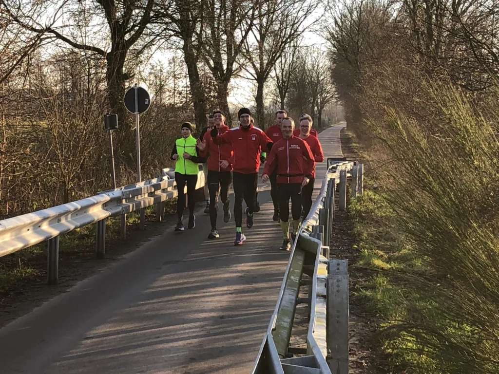 Gruppe von Läufern in Sportkleidung joggt auf einer ländlichen Straße im Winter. Bäume säumen den Weg.