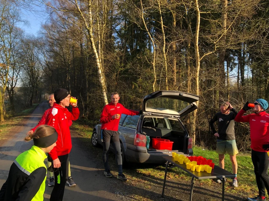 Gruppe von Läufern in rot und schwarz, die an einem sonnigen Morgen an einem Tisch mit Getränken stehen, während sie sich auf einer Waldstraße erfrischen. Ein Auto mit offenem Kofferraum steht im Hintergrund, umgeben von Bäumen.