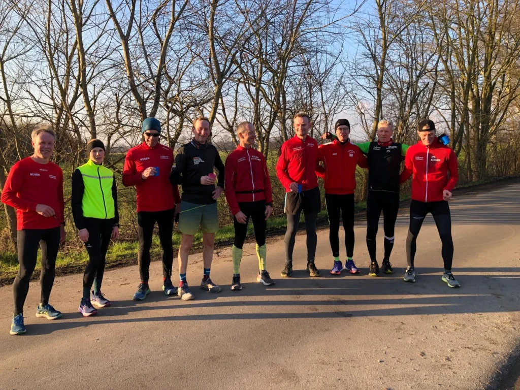 Gruppe von Läufern in roten Trikots, die an einer Straße stehen, umgeben von Bäumen. Sie posieren fröhlich nach dem Training im Freien an einem sonnigen Tag.