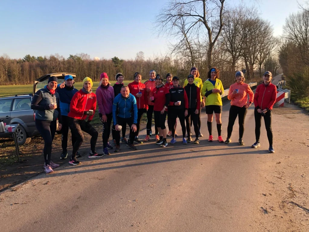 Gruppe von Läufern in bunter Sportkleidung auf ländlicher Straße bei sonnigem Winterwetter, bereit für den Lauf.