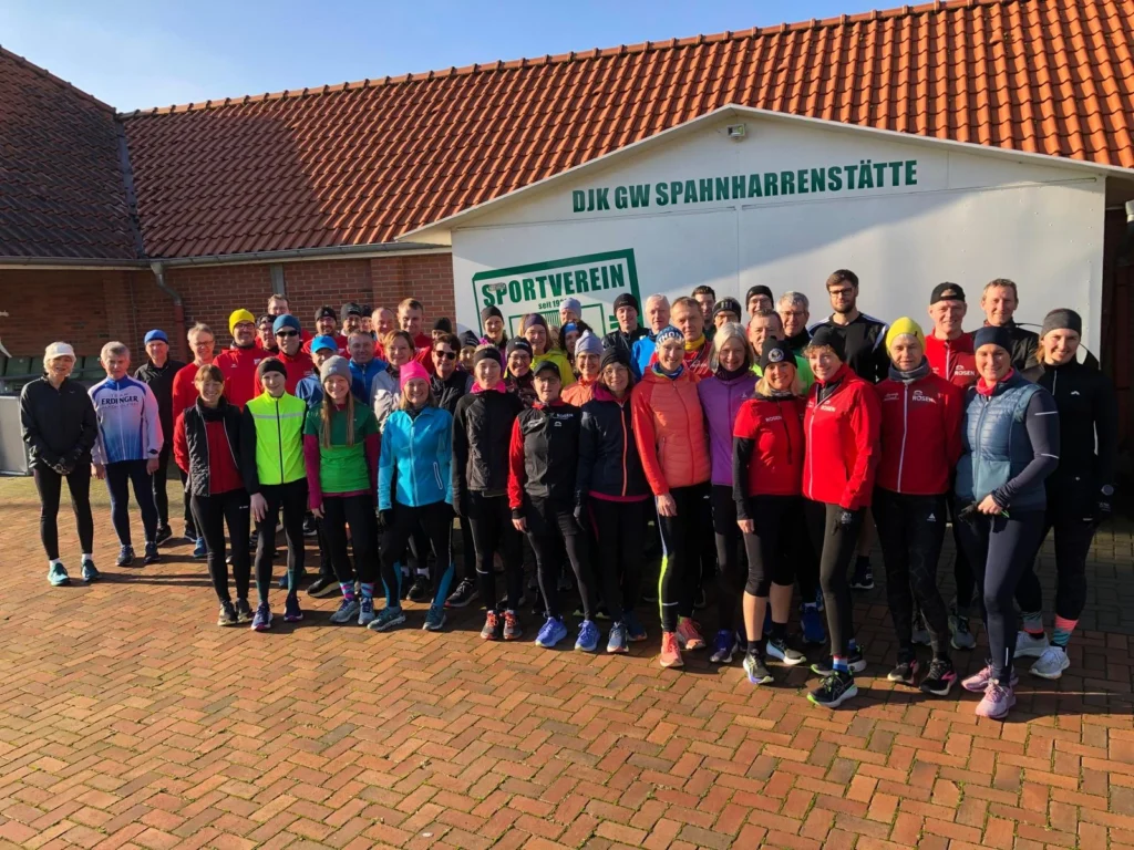 Gruppenfoto von Läufern des DJK GW Spanharrenstätte vor dem Sportverein. Die Teilnehmer sind in Sportkleidung, darunter Jacken und Mützen, und stehen auf einem gepflasterten Platz.