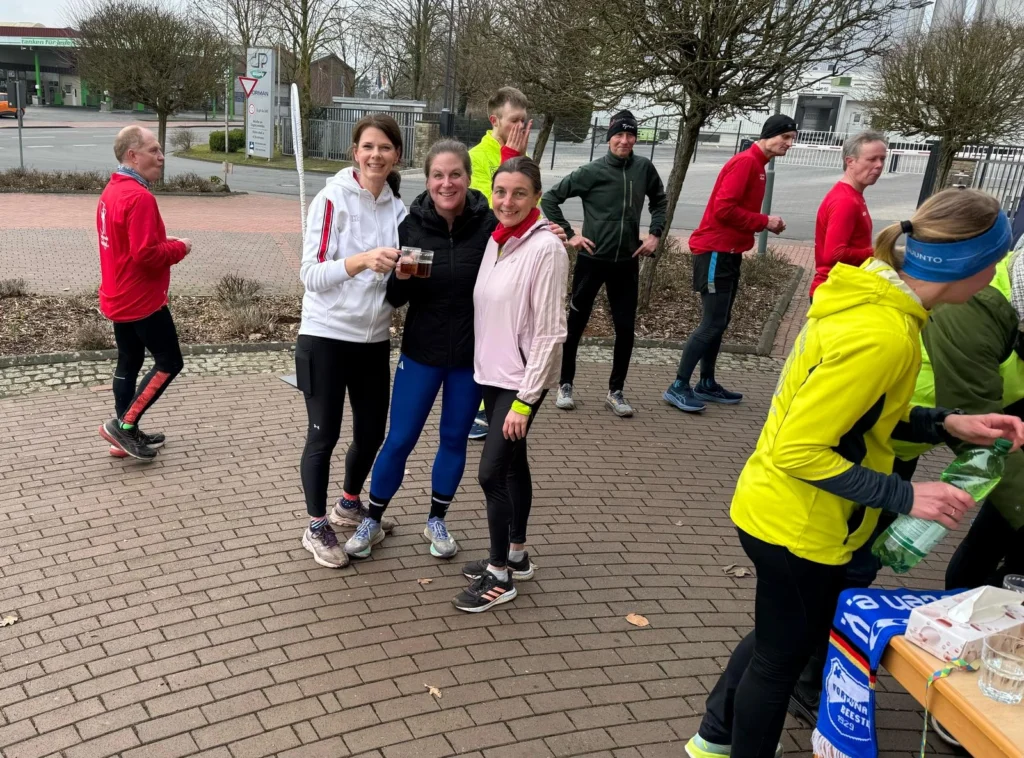 Gruppe von Joggern trifft sich nach einem Lauf, lächelnd und mit Getränken in der Hand auf einem Gehweg.