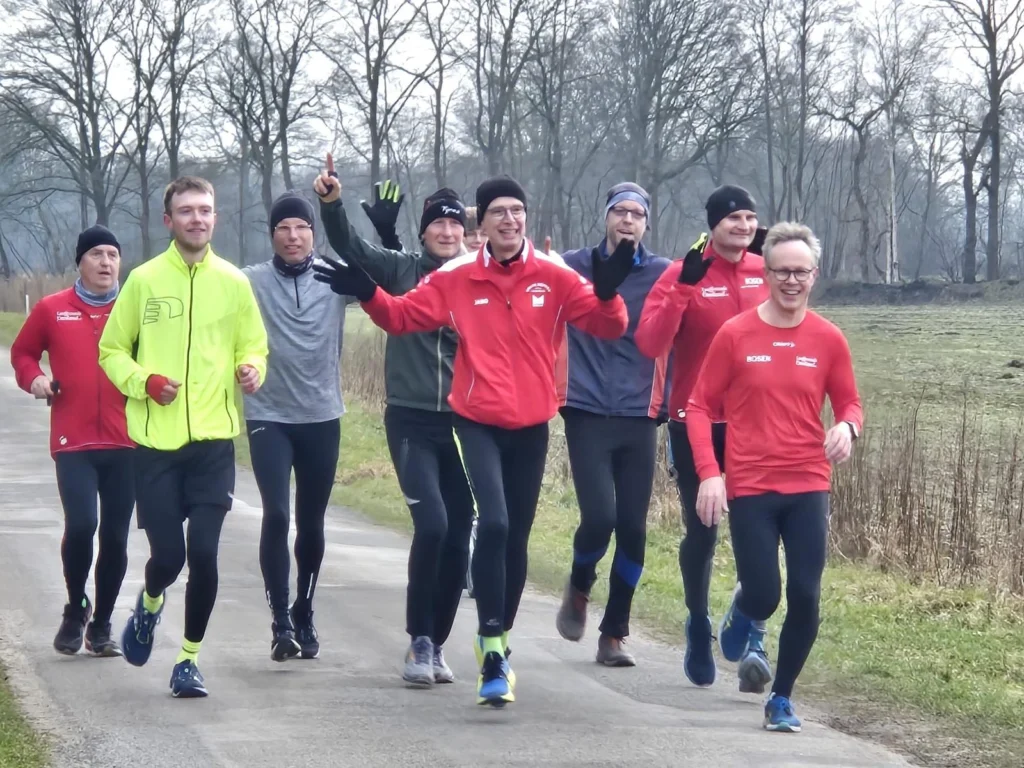 Gruppe von Läufern in sportlicher Kleidung joggt gemeinsam auf einem Weg in der Natur. Sie tragen verschiedene Farben, darunter leuchtendes Gelb und Rot, und scheinen Freude am Laufen zu haben. Bäume und eine grüne Wiese sind im Hintergrund sichtbar.