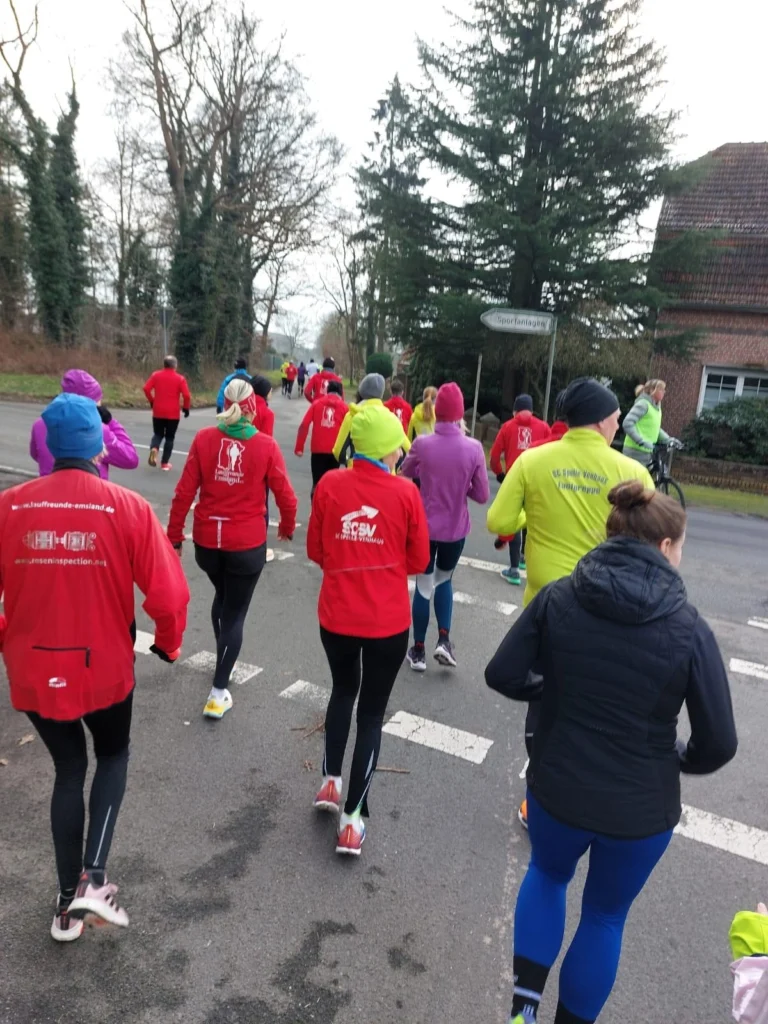 Läufer in bunten Trainingsanzügen joggen auf einer Straße, umgeben von Bäumen und Wohnhäusern. Die Gruppe besteht aus mehreren Teilnehmern in roten, grünen und lila Outfits, die an einem Lauf-Event teilnehmen.