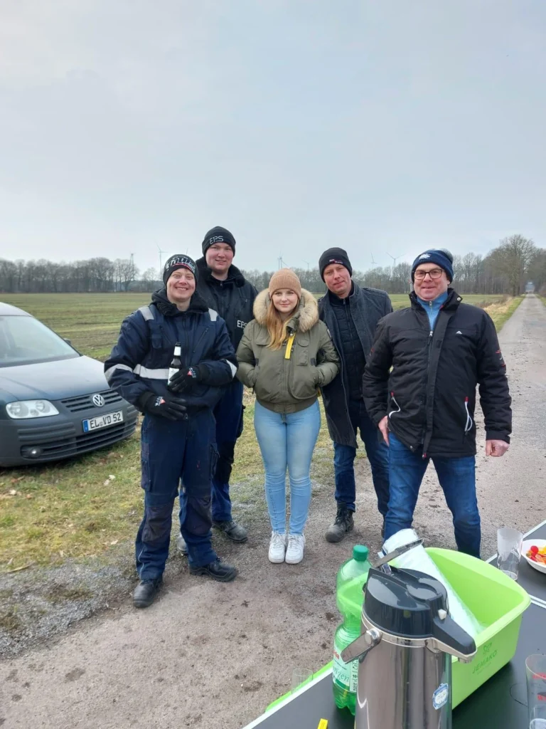 Gruppe von fünf Personen, darunter drei Männer in Arbeitskleidung und zwei Frauen, die an einer Straße stehen. Im Hintergrund sind ein Auto und Windräder sichtbar. Auf einem Tisch im Vordergrund stehen Getränke und Snacks.