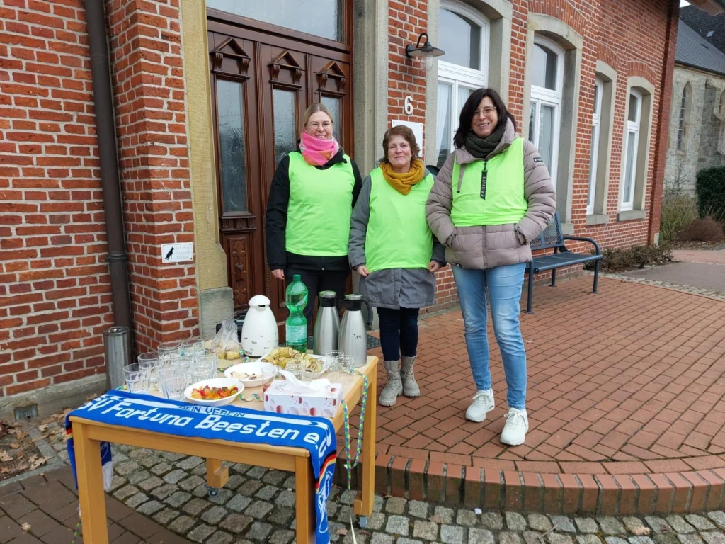 Drei Frauen in leuchtend grünen Westen stehen vor einem Backsteingebäude und lächeln. Vor ihnen befindet sich ein Tisch mit Getränken und Snacks, darunter Gläser, eine Kanne und Süßigkeiten. Im Hintergrund ist eine Bank und ein Weg aus Pflastersteinen sichtbar. Die Szene vermittelt eine freundliche Atmosphäre und zeigt eine Gemeinschaftsveranstaltung.