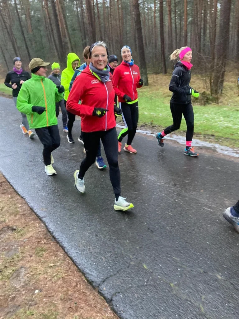 Gruppe von Läufern in sportlicher Kleidung, die auf einem nassen Waldweg joggen. Die Szene zeigt eine Mischung aus verschiedenen Farben und Stilen der Laufbekleidung, während die Teilnehmer motiviert und lächelnd laufen.