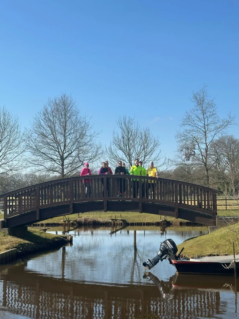 Personengruppe in bunter Kleidung steht auf einer Holzbrücke über einem Fluss bei sonnigem Wetter im Park.