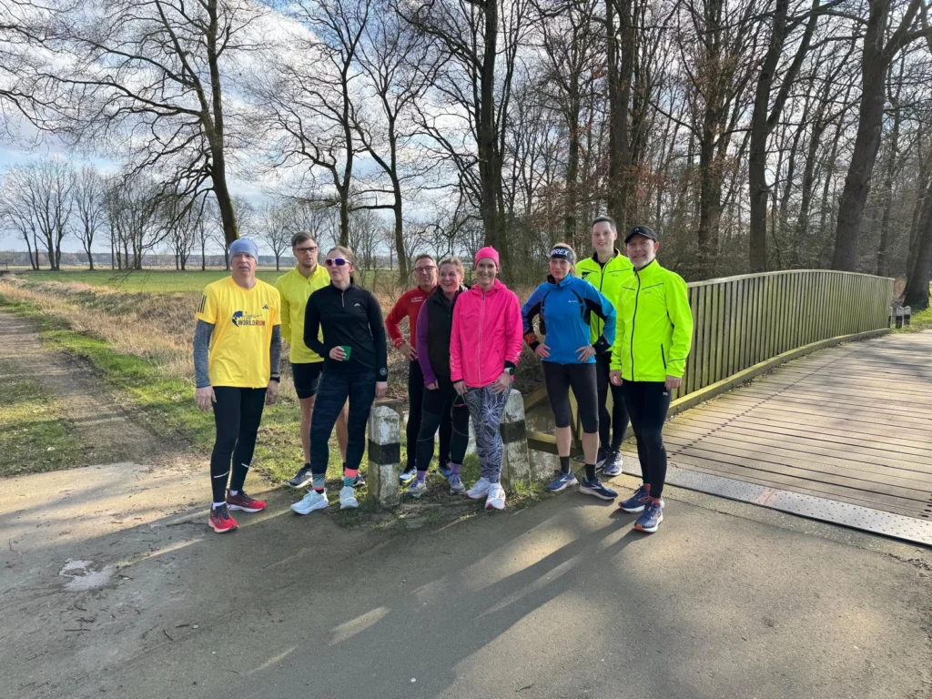 Gruppe von Läufern in bunter Sportkleidung posiert vor einer Brücke im Wald an einem sonnigen Tag für ein Foto.