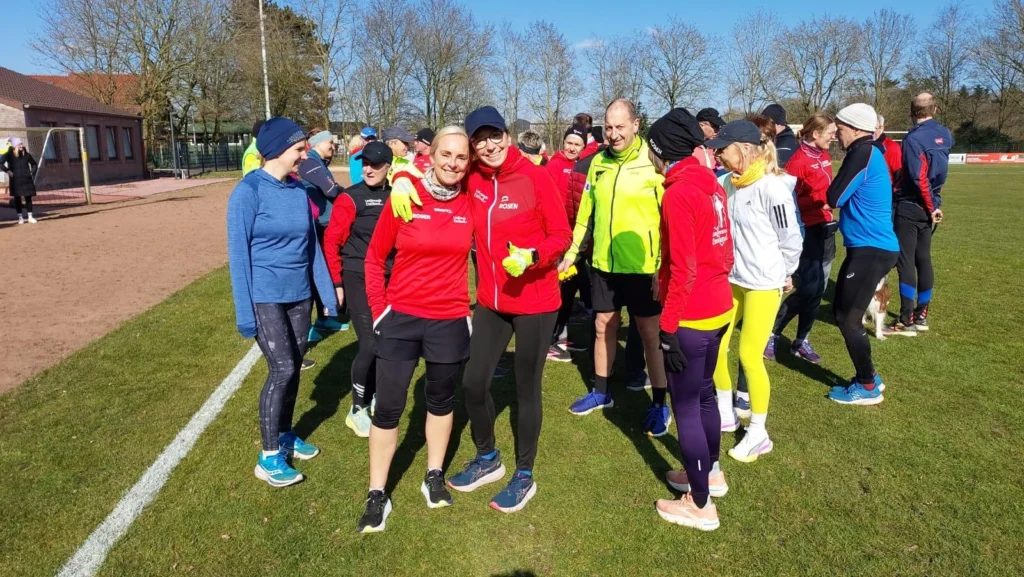 Gruppentraining auf dem Sportplatz mit Teilnehmern in Sportkleidung, darunter lächelnde Frauen in roten Jacken, die sich umarmen. Im Hintergrund sind weitere Sportler und eine klare, sonnige Himmel zu sehen. Ideal für Lauf-Community-Events und sportliche Aktivitäten.