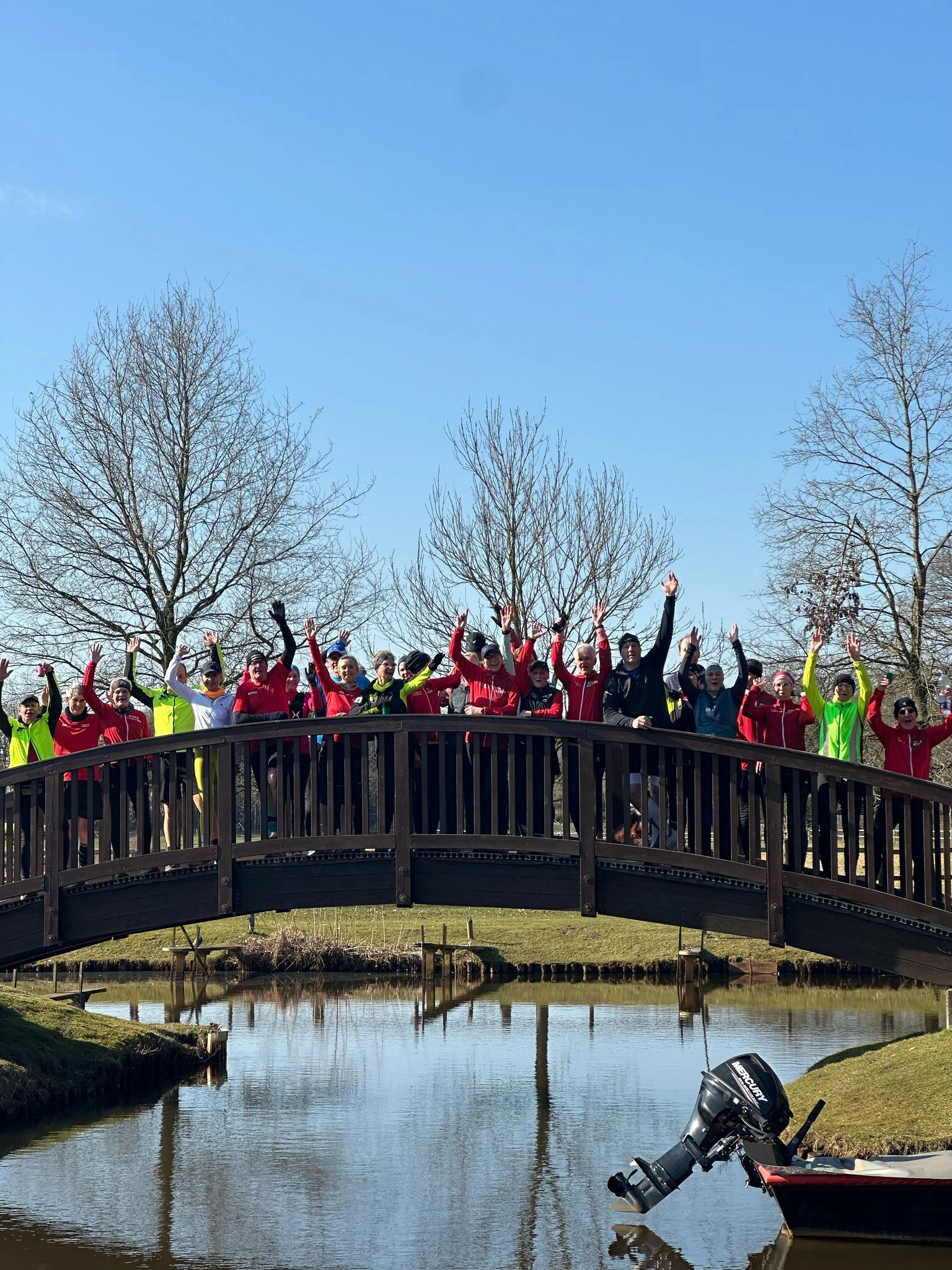 Gruppe fröhlicher Menschen steht jubelnd auf einer Holzbrücke über einem Teich an einem sonnigen Tag im Park.