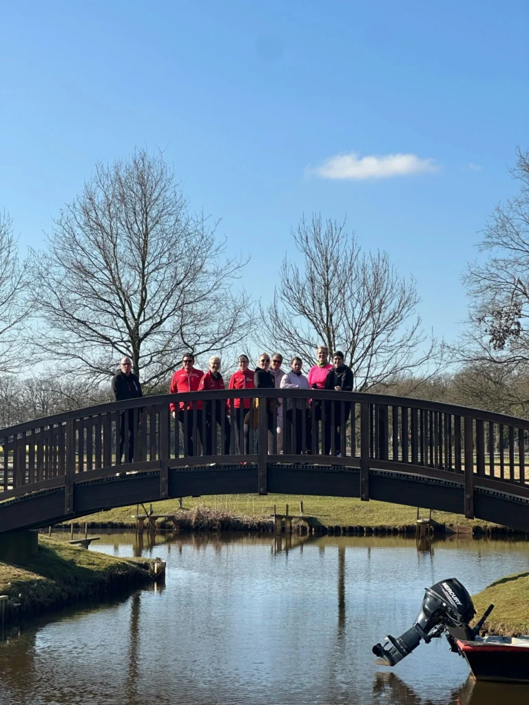 Gruppe von Menschen auf einer Holzbrücke über einem ruhigen Teich an einem sonnigen Tag, umgeben von kahlen Bäumen und einem Boot mit Außenbordmotor im Vordergrund.