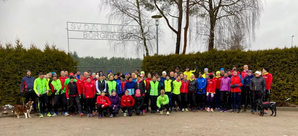 Gruppe von Läufern in bunter Sportbekleidung posiert bei einem Lauf-Event im Freien vor einem Schild und Bäumen.