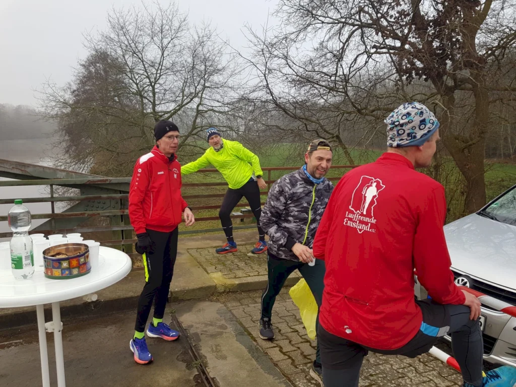 Gruppe von Läufern in sportlicher Kleidung, die sich an einem Flussufer treffen. Im Vordergrund stehen ein Tisch mit Getränken und Snacks sowie eine Wasserflasche. Die Umgebung ist neblig und zeigt Bäume im Hintergrund.