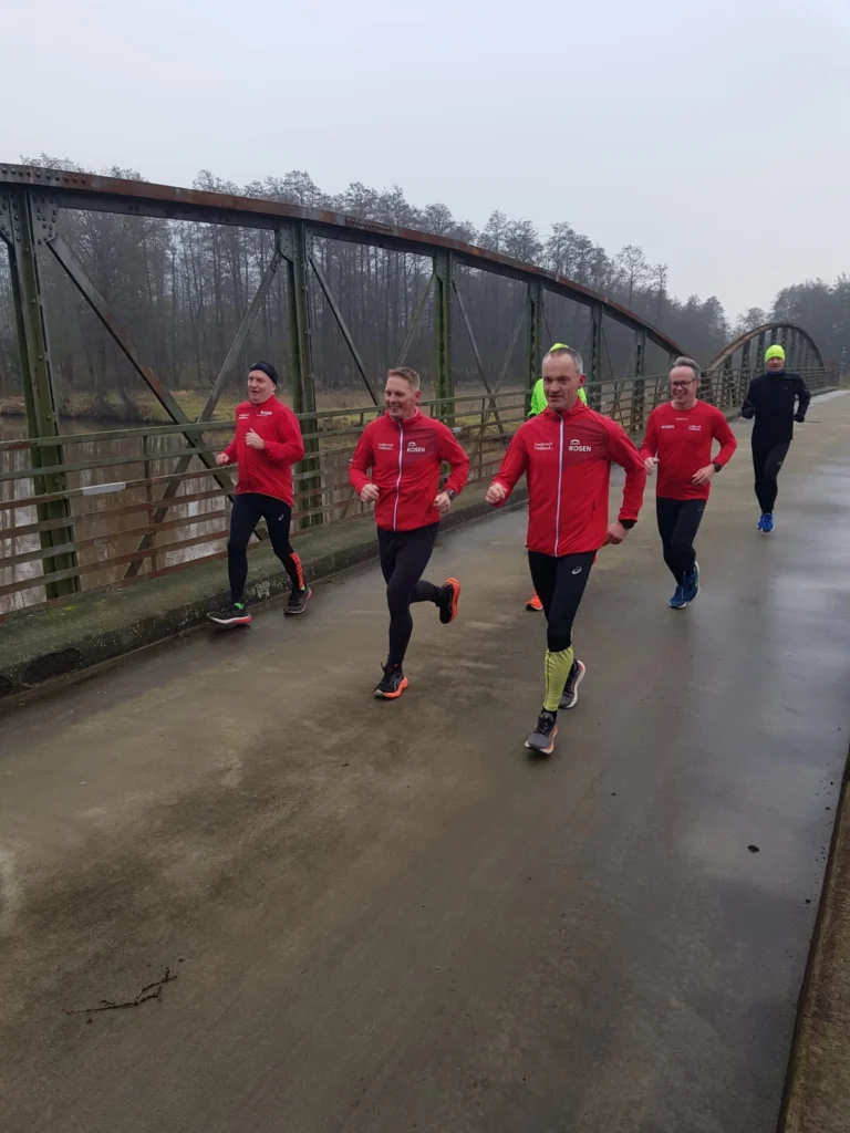 Gruppe von Läufern in roten Jacken joggt auf einer Brücke in der Natur, umgeben von Bäumen und Nebel. Ideal für Sport- und Fitnessinhalte.