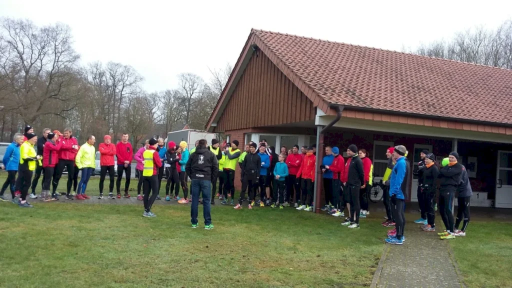 Gruppentraining von Läufern in sportlicher Kleidung vor einem Vereinsgebäude, während ein Trainer Anweisungen gibt. Die Teilnehmer sind in verschiedenen Farben gekleidet und stehen auf einer grünen Wiese.