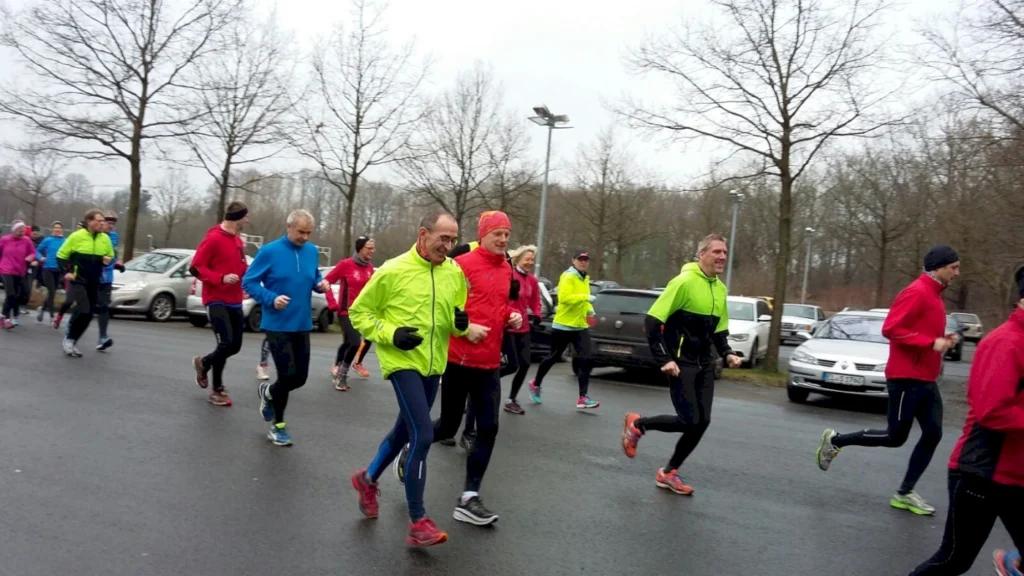 Läufer in bunten Sportoutfits joggen auf einem Parkplatz, umgeben von kahlen Bäumen und Autos, bei bewölktem Wetter.