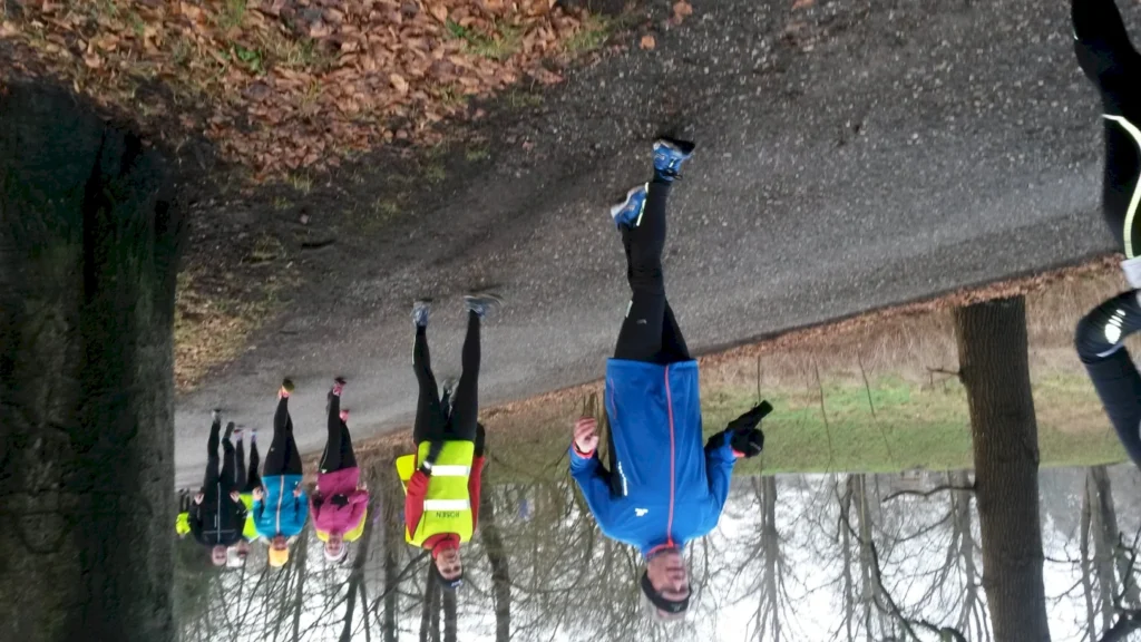 Gruppe von Sportlern beim Laufen auf einem Waldweg, einige tragen reflektierende Kleidung, um Sicherheit zu gewährleisten. Herbstliche Landschaft mit Laub und Bäumen im Hintergrund.
