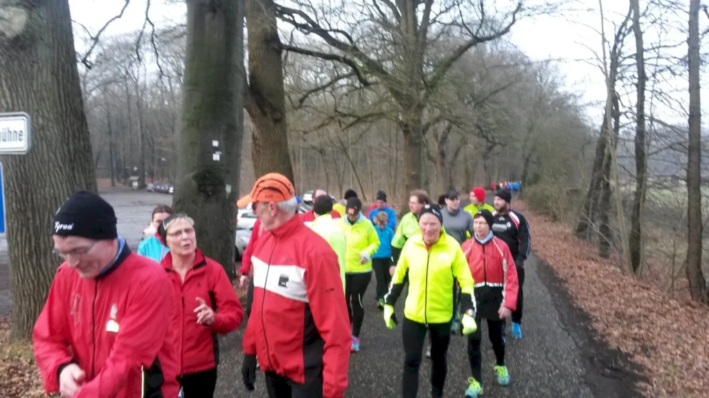Gruppierung von Läufern in bunten Sportjacken, die auf einem Waldweg joggen. Der Hintergrund zeigt Bäume und eine Straße, während die Teilnehmer aktiv an einem Lauf-Event teilnehmen.