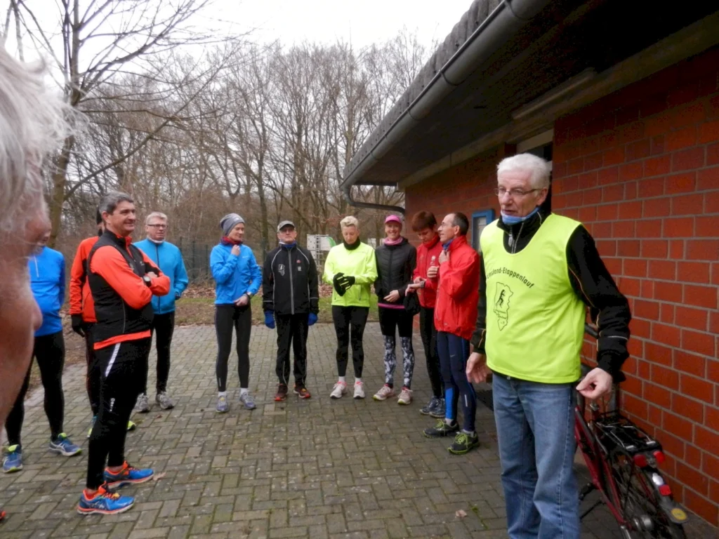 Gruppe von Läufern in Sportkleidung, die sich vor einem Gebäude versammeln. Ein älterer Mann in einem gelben Laufshirt erklärt etwas. Die Szene spielt sich in einer natürlichen Umgebung mit Bäumen im Hintergrund ab, ideal für ein Lauftraining oder eine Sportveranstaltung.