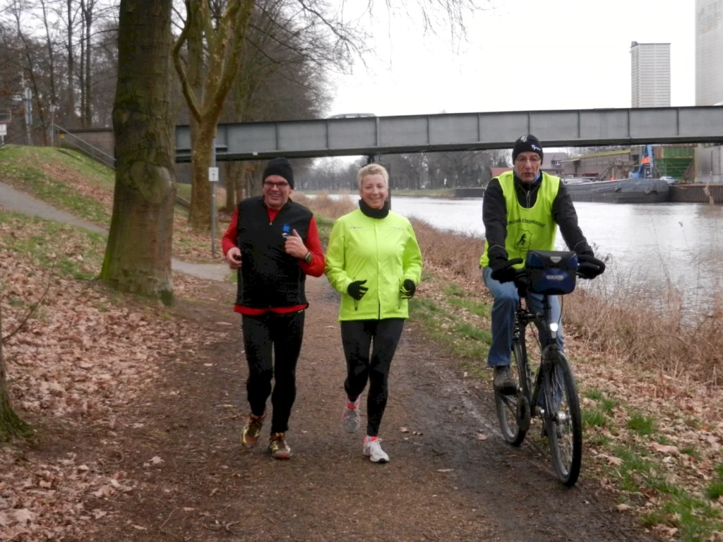 Drei Personen, zwei Jogger und ein Radfahrer, bewegen sich entlang eines Weges am Wasser. Die Jogger tragen sportliche Kleidung, darunter eine leuchtend gelbe Jacke, während der Radfahrer einen grünen Sicherheitswesten trägt. Im Hintergrund sind Bäume und ein Brücke sichtbar, was eine aktive Freizeitgestaltung in der Natur zeigt.