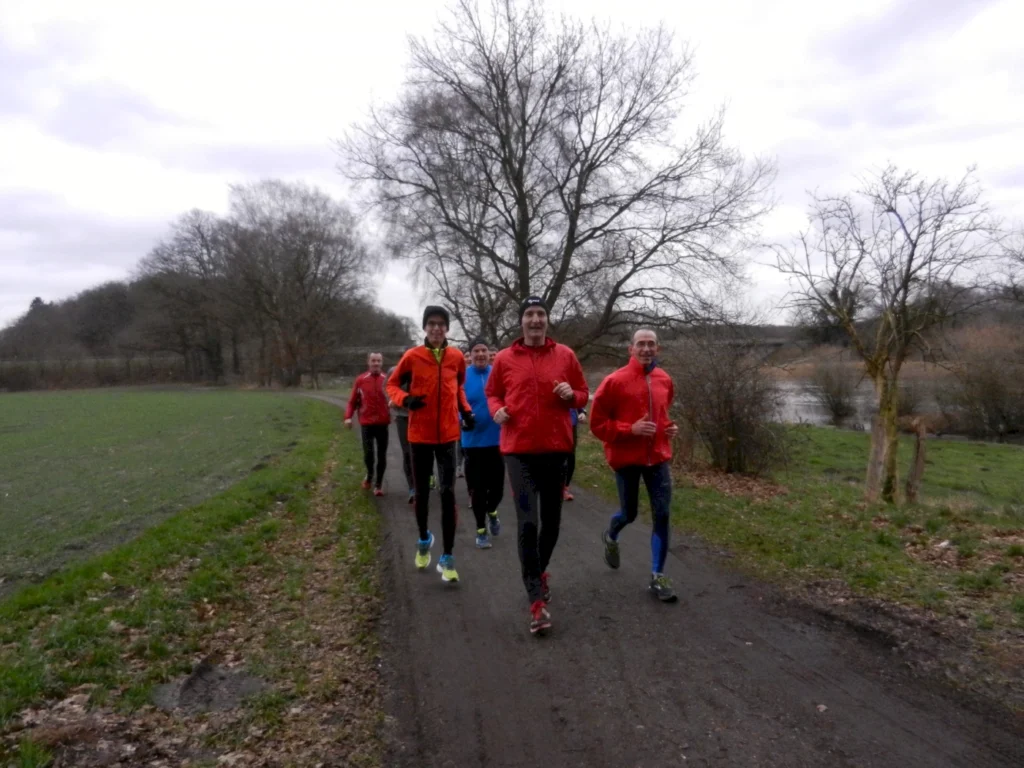 Gruppe von sechs Läufern in sportlicher Kleidung, die auf einem malerischen Weg entlang eines Flusses joggen, umgeben von Bäumen und grünem Gras. Ideal für Fitness und Outdoor-Aktivitäten.