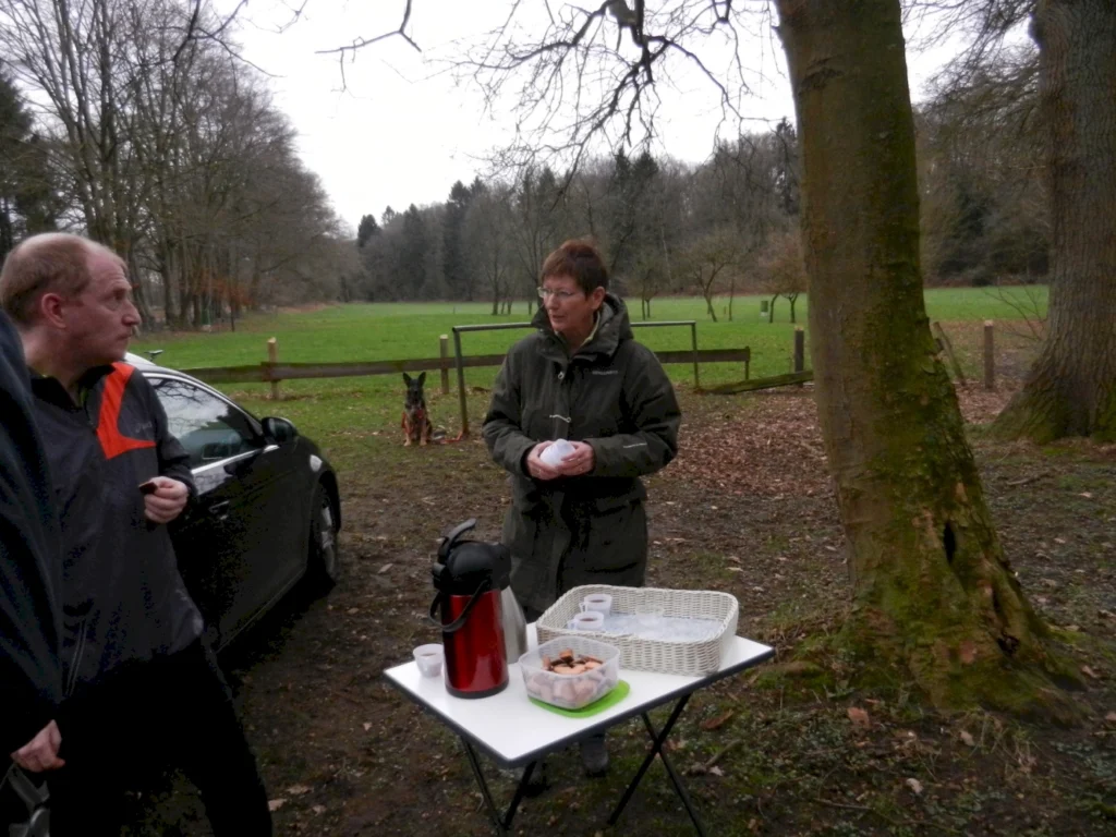 Zwei Personen stehen an einem Tisch im Freien, der mit Snacks und einer Thermoskanne gedeckt ist. Im Hintergrund ist ein Hund und eine grüne Wiese zu sehen, umgeben von Bäumen. Die Szene vermittelt eine entspannte Atmosphäre in der Natur.