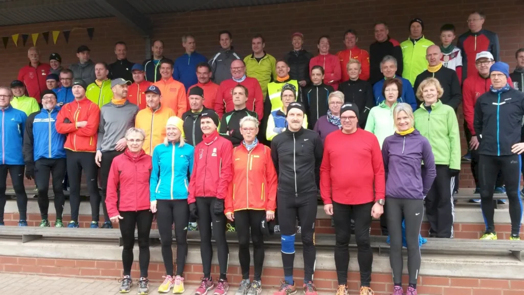 Gruppenfoto von Laufbegeisterten in bunten Sportjacken und -hosen, die auf Treppen stehen. Die Teilnehmer sind sowohl Männer als auch Frauen unterschiedlichen Alters und posieren für ein gemeinsames Bild. Die Veranstaltung findet wahrscheinlich im Freien statt, bei kühlem Wetter.