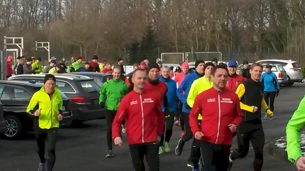 Gruppensport-Event mit zahlreichen Läufern in bunten Sportjacken, die auf einem Parkplatz vor einem Sportgelände joggen. Im Hintergrund sind Autos und Bäume zu sehen.