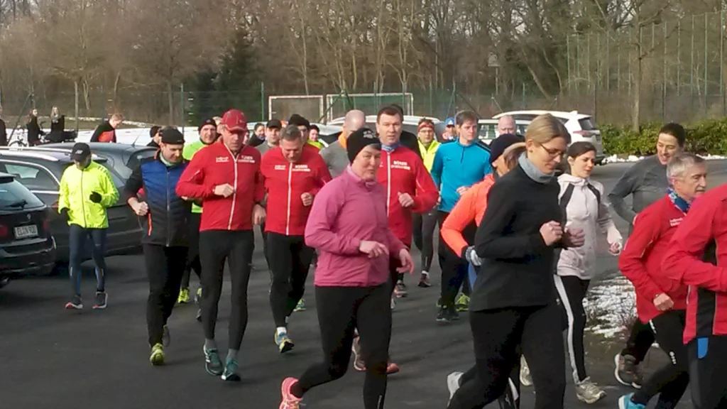 Gruppe von Läufern in sportlicher Kleidung, die auf einem Parkplatz joggen. Im Hintergrund sind Bäume und ein Sportplatz sichtbar. Das Bild zeigt eine Vielzahl von Personen unterschiedlichen Alters, die an einem Lauf-Event teilnehmen.