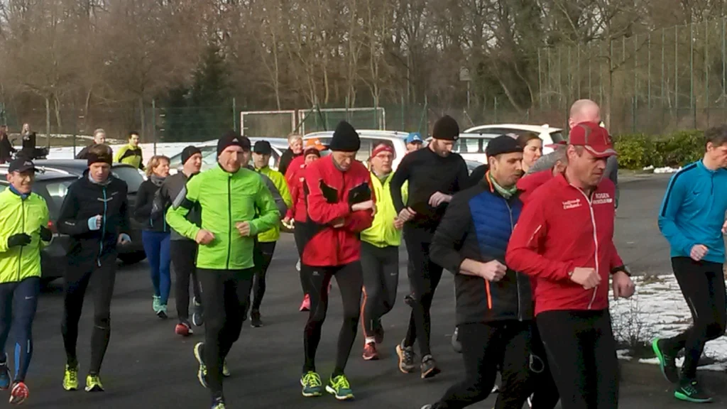 Gruppentraining von Läufern in sportlicher Kleidung, die auf einem Parkplatz joggen. Im Hintergrund sind Autos und eine Sportanlage sichtbar.