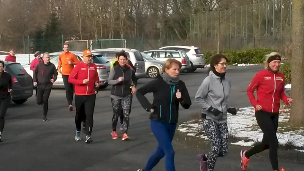 Gruppe von Menschen beim Laufen auf einem Parkplatz, umgeben von Autos und Bäumen, im Winter mit leichtem Schnee. Die Teilnehmer tragen sportliche Kleidung und zeigen Freude am Laufen.