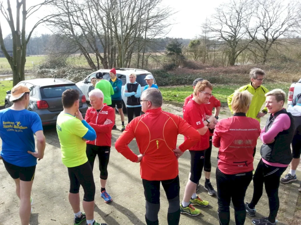 Gruppe von Läufern in Sportkleidung, die sich vor einem Training austauschen, umgeben von Autos und einer natürlichen Landschaft im Hintergrund. Ideal für die Förderung von Laufgemeinschaften und Fitnessveranstaltungen.